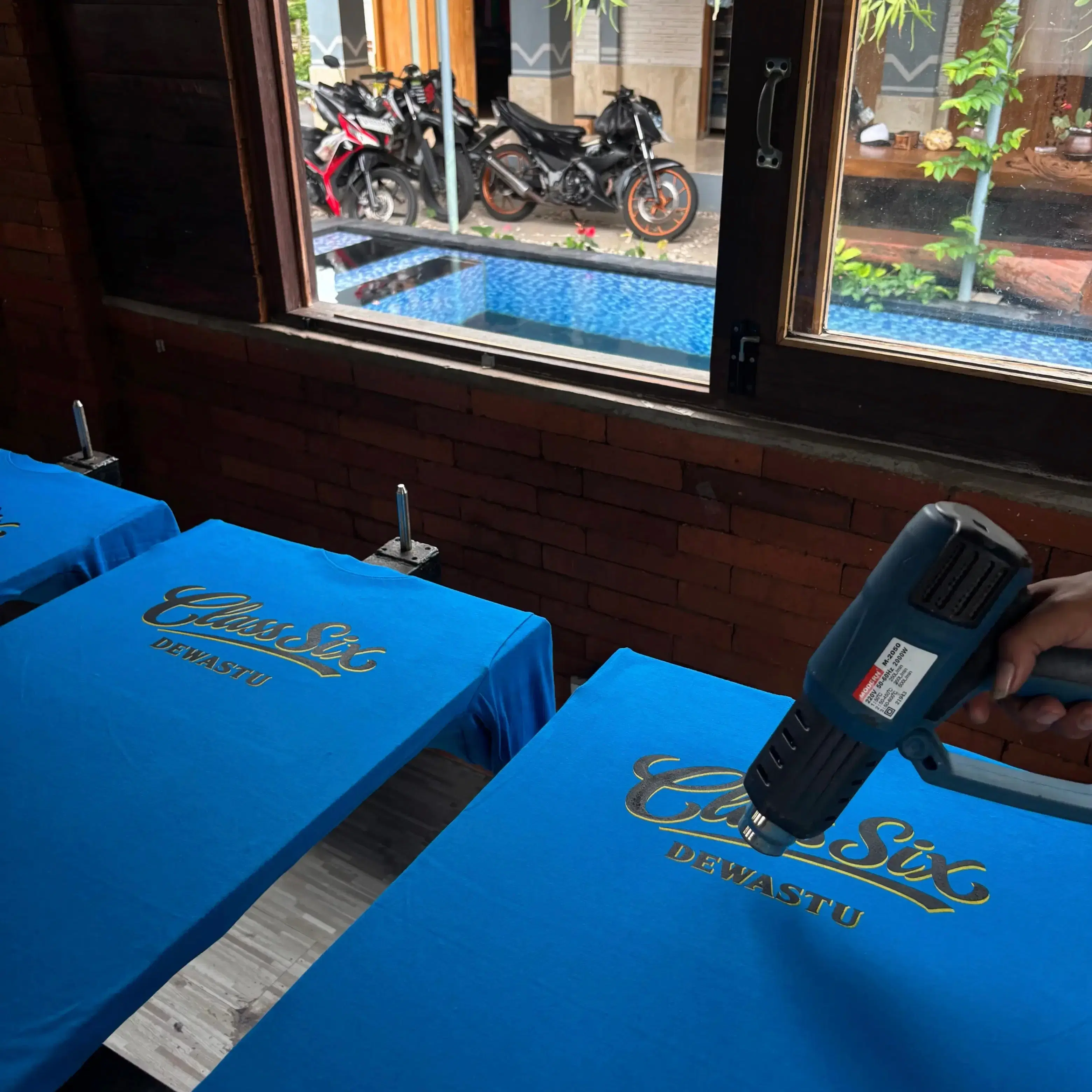 Proses heat press kaos custom Class Six Dewastu di workshop Blumbang ID, Jeto Klaten — kolam renang workshop terlihat dari jendela