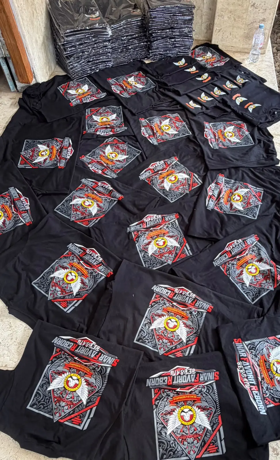 Puluhan kaos hitam Sinar Favorit Reborn berjejer di lantai, sablon plastisol empat warna merah putih abu-abu, logo lingkaran dua tangan bergandengan diapit sayap dalam bingkai gunungan berornamen ukiran, sebagian masih terbungkus plastik di bagian belakang
